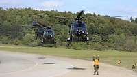 Vier Hubschrauber im Landeanflug. Ein Soldat mit Warnweste weist sie ein.