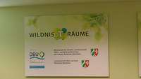 Ein Schild an einer Wand, welches die Aufschrift Wildnis(t)räume trägt.