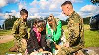 Zwei Soldaten knien mit 2 Frauen auf einer freien Wiese am Boden. 