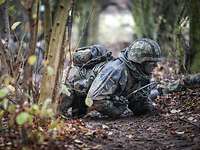 Ein Soldat kriecht durch das Gelände. Weitere Soldaten unscharf im Hintergrund.