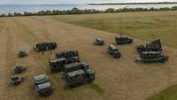 Mehrere Militärfahrzeuge stehen auf einem Feld. Das Feld grenzt im Hintergrund ans Meer.