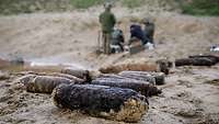 Alte Munition liegt aufgereiht auf Sandboden, im Hintergrund Soldaten.