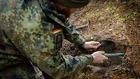 Ein Soldat kniet und gräbt vorsichtig im Waldboden.