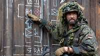 Ein Soldat kniet vor einer Holzwand, auf der Symbole mit Kreide stehen. Er weist darauf.