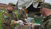 Der Kompaniefeldwebel gibt aus einer rechteckigen Therme an Soldaten eine heiße Suppe aus.