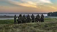 Eine Gruppe Soldaten steht kurz vor Sonnenaufgang auf einem Feld vor einem nebligen Hintergrund.