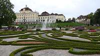 Der prunkvolle Schlossgarten mit Brunnen und bunten Blumen vor einem Schloss.