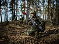 Ein Soldat kniet mit Waffe im Wald. Im Hintergrund laufen Soldaten mit roten Flaggen.