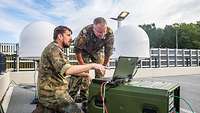 Zwei Soldaten schauen auf einen Laptop. Im Hintergrund sind zwei Kugelförmige Antennen zu sehen.