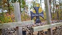 Das Eiserne Kreuz mit Text „Den Toten zur Ehr“ und davor Holzkreuze mit Namen im Wald der Erinnerung