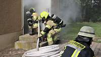 Brandschützer in Feuerwehruniform gehen mit einem Schlauch durch die Tür eines rauchenden Gebäudes.