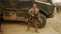 Ein Bundeswehrsoldat in Wüstentarnuniform mit Waffe steht vor einem Radfahrzeug Dingo im Sand.