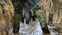 Soldaten laufen in grünen Gummistiefeln durch einen mit Wasser gefluteten braunen Erdgraben.