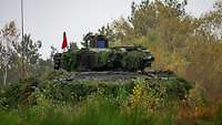 Ein Schützenpanzer Puma steht mit einer roten Flagge zwischen grünem Buschwerk in Stellung.