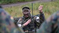 Ein Soldat mit Funkgerät und weißer Armbinde spricht auf einer Lichtung vor einer Gruppe Soldaten.