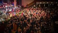 Großer Ballsaal mit Bühne und Tanzfläche, auf der viele Paare tanzen. Im Hintergrund und auf der Empore Tische.