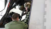 Ein junger Mann sitzt in einem Cockpit eines Hubschraubers, ein Mann in Uniform erklärt ihm alles.