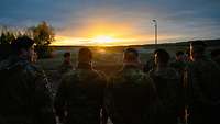 Soldaten stehen mit Blick auf den Übungsplatz. Am Horizont geht die Sonne auf.
