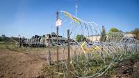 A barbed wire fence. In the background there are Tents and buildings.