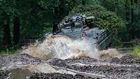 Ein Schützenpanzer Marder fährt im Wald auf einem schlammigen Weg, Wasser spritzt auf.