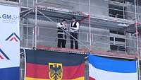 Zwei Männer in Zimmermannskluft stehen auf einem Baugerüst, am Gerüst hängen farbige Flaggen
