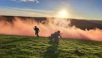 Zwei Soldaten rennen vor einer Nebelwand weg. Die tiefstehende Sonne färbt den Nebel orange. 