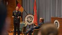 Konteradmiral Ralf Kuchler steht auf der Bühne, daneben sitzt der Verteidigungsminister