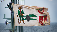 Eine beige handgemalte Flagge mit einem Jäger, einem Wildschwein und zwei Wappen.