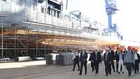 A group of people in a dock hall walking past a ship with scaffolding.