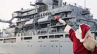 Vor einem Schiff steht eine Person in einem Weihnachtsmannkostüm, die den Soldaten darauf zuwinkt
