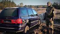 Ein Auto mit einem angeklebten Sprengsatz steht auf einem Gelände, daneben steht ein Soldat.