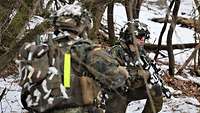 Ein Soldat mit leichter Schneetarnung überwacht das Vorfeld. Ein anderer läuft auf ihn von der Seite zu.