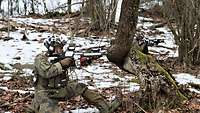 Zwei Soldaten hocken mit Gewehren im Wald. An ihren linken Armen tragen sie rote Armbinden.
