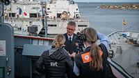 Ein Soldat wird von einem Fernsehteam interviewt, im Hintergrund mehrere Schiffe im Hafen. 