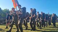 Mehrere Soldatinnen und Soldaten laufen mit Gepäck über eine Wiese. Einer von ihnen trägt eine Fahne.