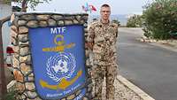Ein Soldat steht neben dem Wappen der MTF, welches ihm bis zur Schulter reicht.