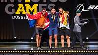 Drei Sportler mit Flaggen und Medaillen stehen jubelnd auf der Bühne bei den Invictus Games 2023
