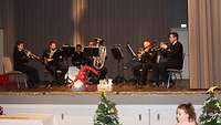 Sechs Musiker in Marineuniform spielen Blasinstrumente auf einer Bühne im Saal.