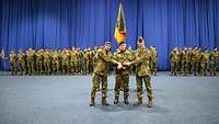 Drei Soldaten stehen nebeneinander und schütteln sich die Hände. Im Hintergrund stehen Soldatinnen und Soldaten in Formation.