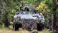 Zwei Soldaten sitzen auf einem Schützenpanzer, der auf einem Waldweg fährt.