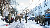 Mehrere Bundeswehrangehörige schieben mit Schneeschiebern den Schnee von einer Straße.