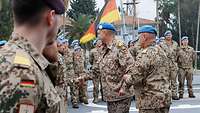 Ein Soldat in Uniform mit blauem Barett beglückwünscht einen Soldaten. Weitere Soldaten stehen angetreten auf dem Oberdeck.