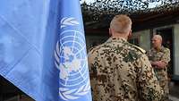 Im Vordergrund sieht man die UN-Flagge. Im Hintergrund sieht man zwei Soldaten in Uniform.
