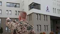 Ein Soldat in Uniform steht vor einem Gebäude und zeigt mit einem Finger auf dieses.