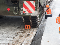 Grüne Kettenfahrzeuge stehen auf Bahnwaggons und Personen in orangenen Warnjacken kontrollieren diese.