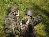 Zwei Soldaten mit Helm und Schutzweste stehen vor einem Wald. 