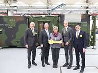 A woman is holding a big key and is standing with four men outside a camouflaged container.