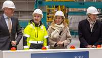 Two women and two men in white protective helmets behind a board with red switches.