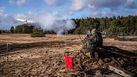 Auf einem Feld schießen drei kniende Soldaten mit einer Panzerabwehrwaffe. Rauch entsteht.