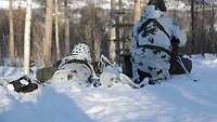 Zwei Soldaten in weißer Uniform liegen im Schnee. Vor ihnen ist ein Wald.